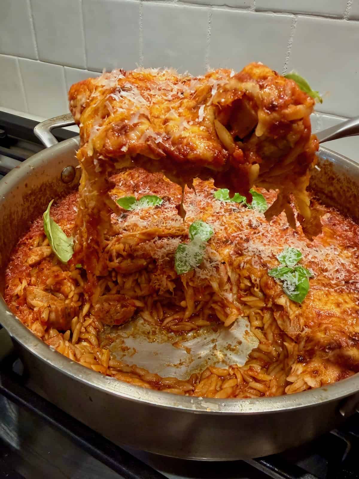 A spoon lifts cheesy sausage orzo from the pan with melted cheese stretching.