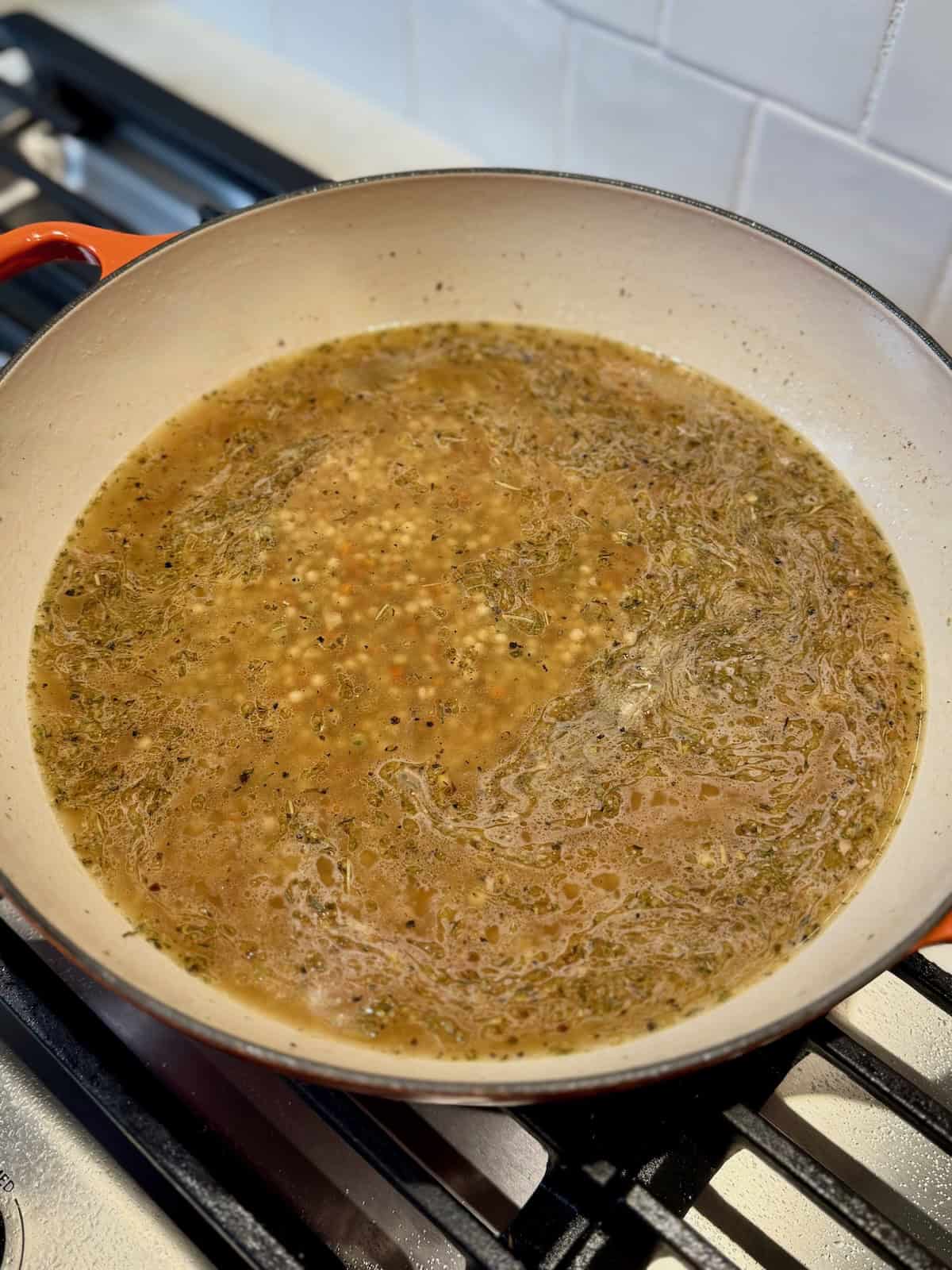 Pearl couscous cooking in broth before adding chicken thighs back to the pan.