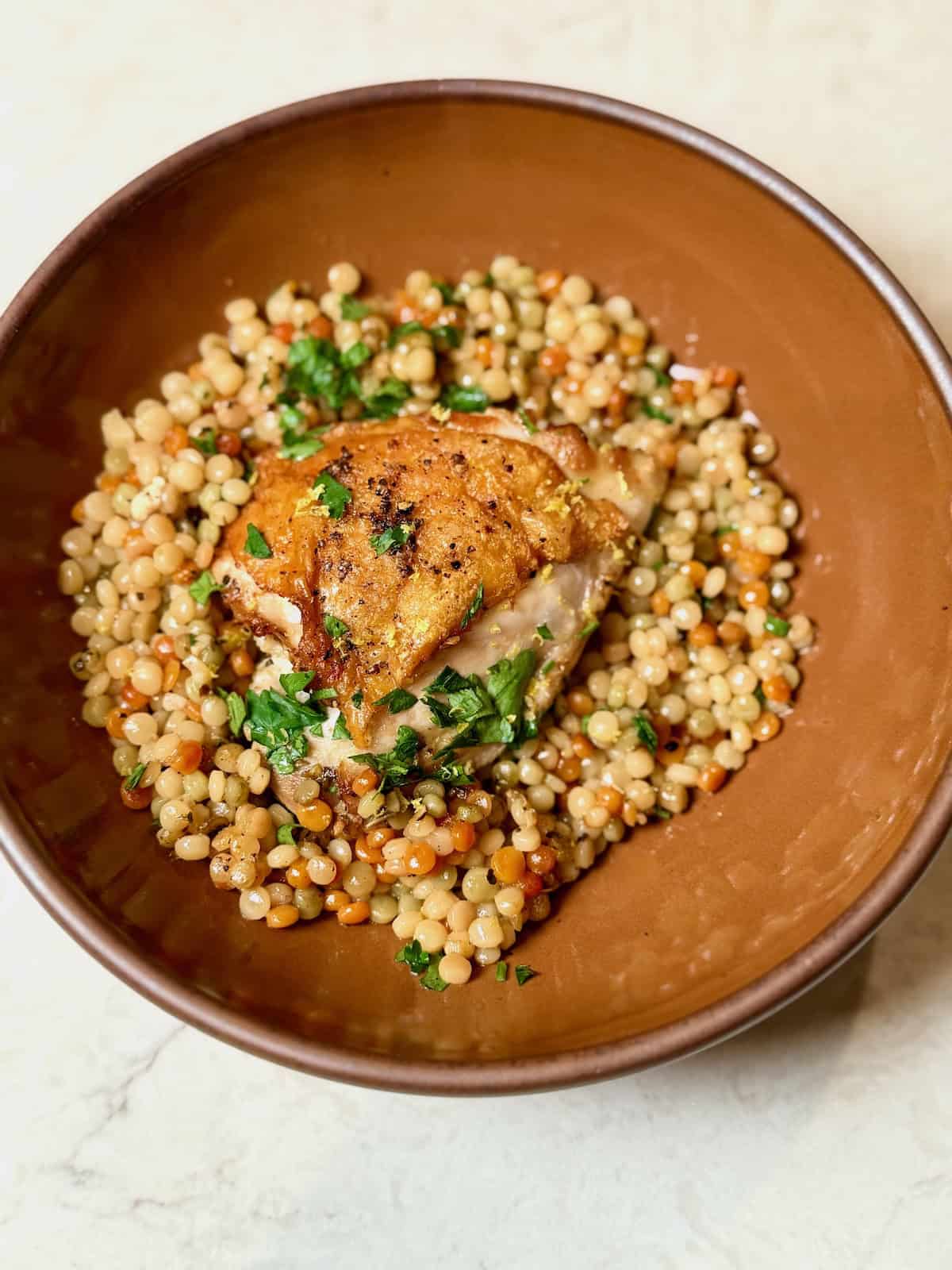 A serving of crispy chicken thighs with lemon couscous in a brown bowl.
