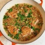 An orange braiser with crispy chicken thighs and lemon couscous sprinkled with fresh parsley.