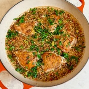 An orange braiser with crispy chicken thighs and lemon couscous sprinkled with fresh parsley.