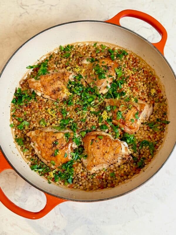 An orange braiser with crispy chicken thighs and lemon couscous sprinkled with fresh parsley.