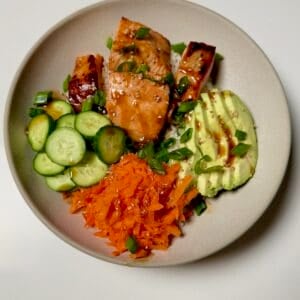 A teriyaki salmon bowl with rice, avocado, cucumber, shredded carrots, and cooked salmon arranged in sections.