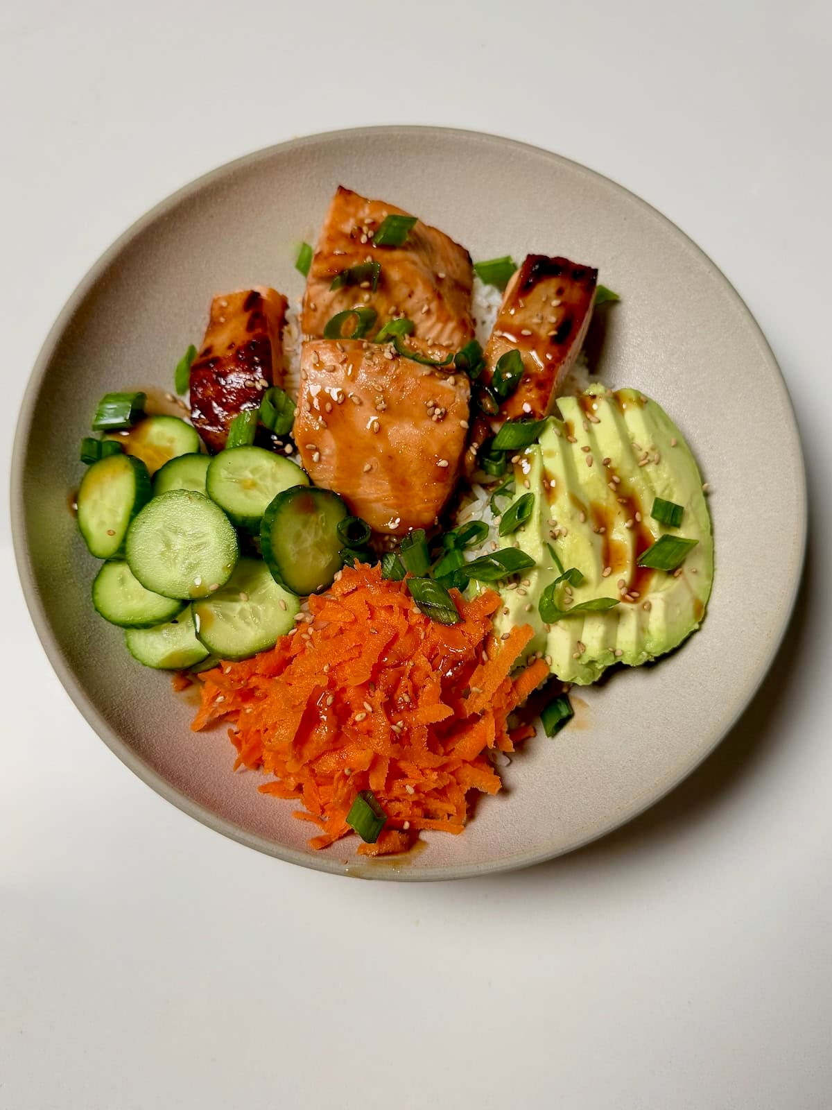 A teriyaki salmon bowl with rice, avocado, cucumber, shredded carrots, and cooked salmon arranged in sections.