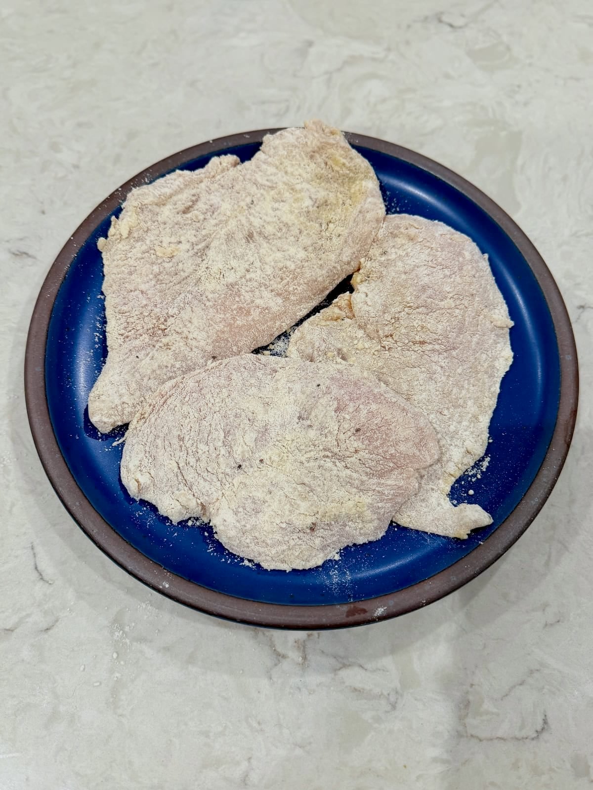 Thin chicken cutlets are evenly coated in seasoned flour on a plate.