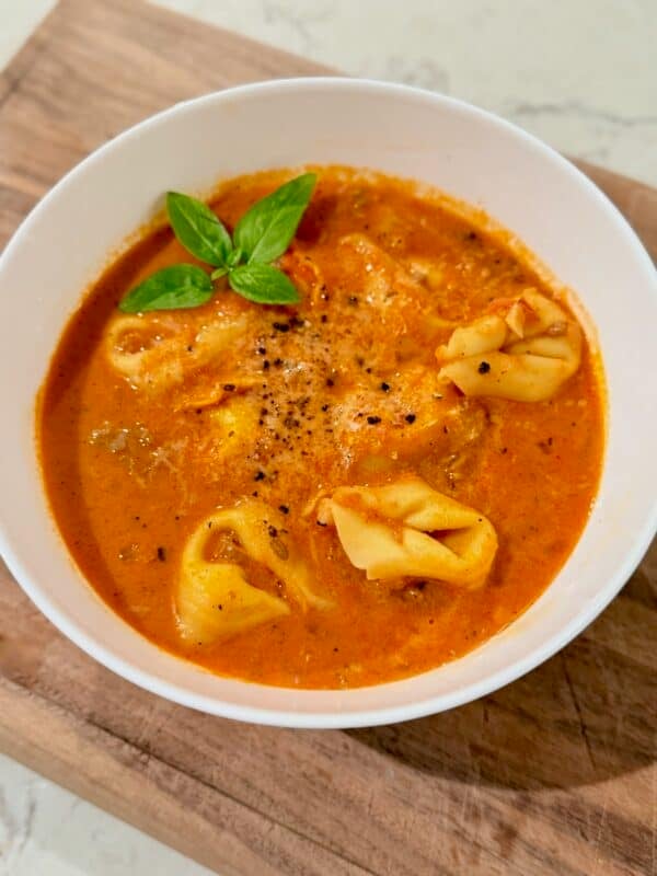 A white bowl of creamy tomato tortellini soup sits on a wooden board with fresh basil.