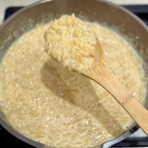 A wooden spoon holds a scoop of creamy orzo mac and cheese over the skillet.