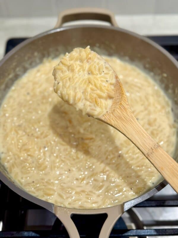 A wooden spoon holds a scoop of creamy orzo mac and cheese over the skillet.
