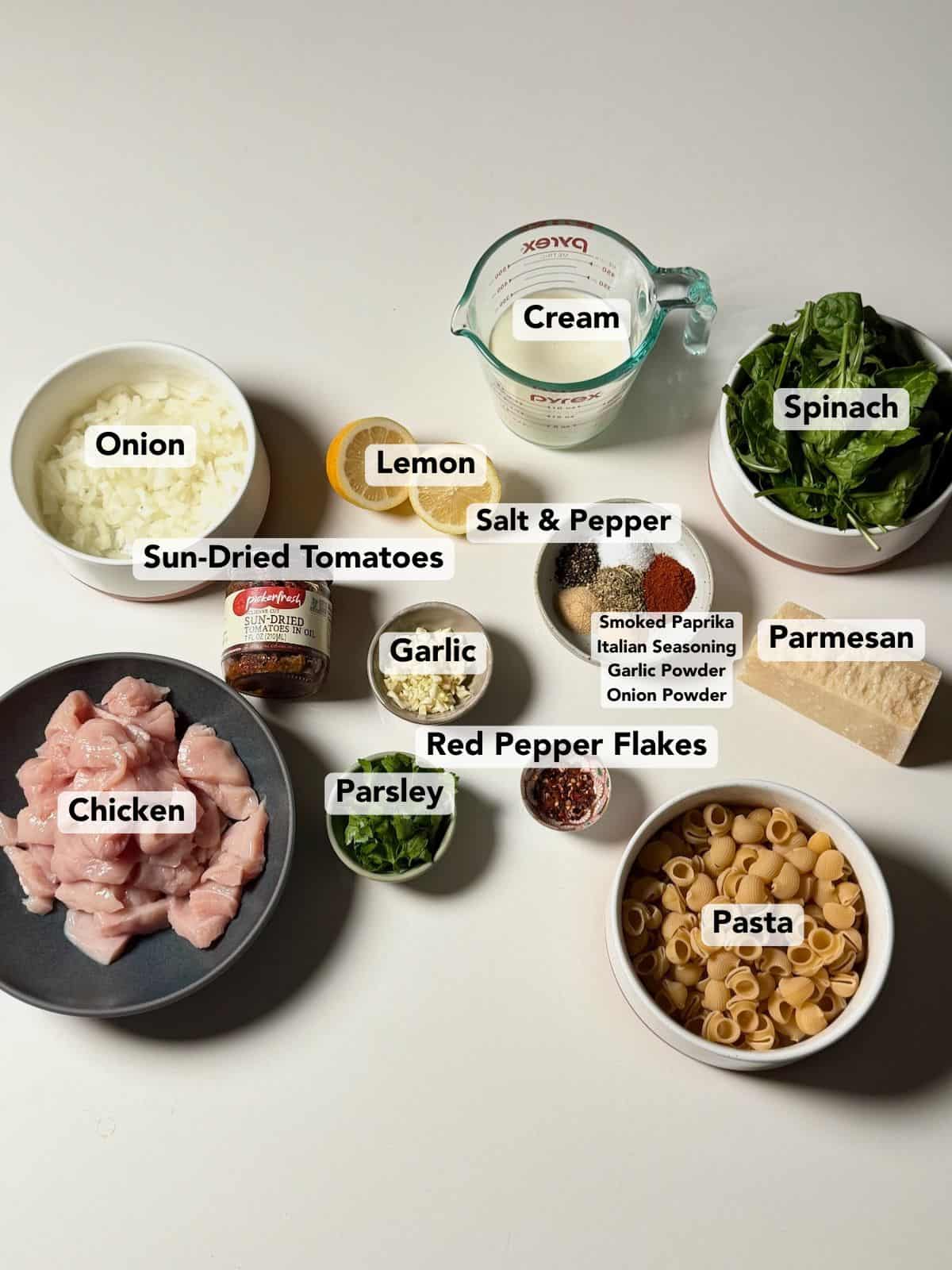 Ingredients for creamy sun-dried tomato chicken pasta arranged on a countertop, including chicken, pasta, sun-dried tomatoes, cream, spinach, and seasonings.