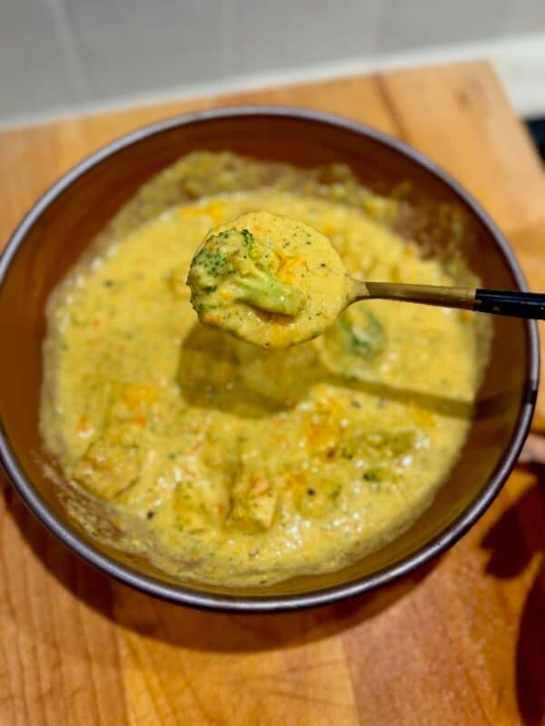 A spoon holds a thick, creamy spoonful of chicken broccoli cheddar soup with a broccoli floret, above a brown bowl of soup in the background.