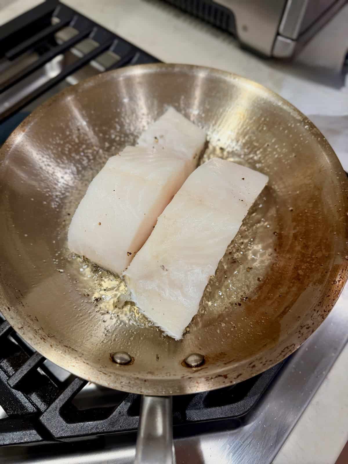 Two Chilean sea bass fillets cook skin-side down in a pan.