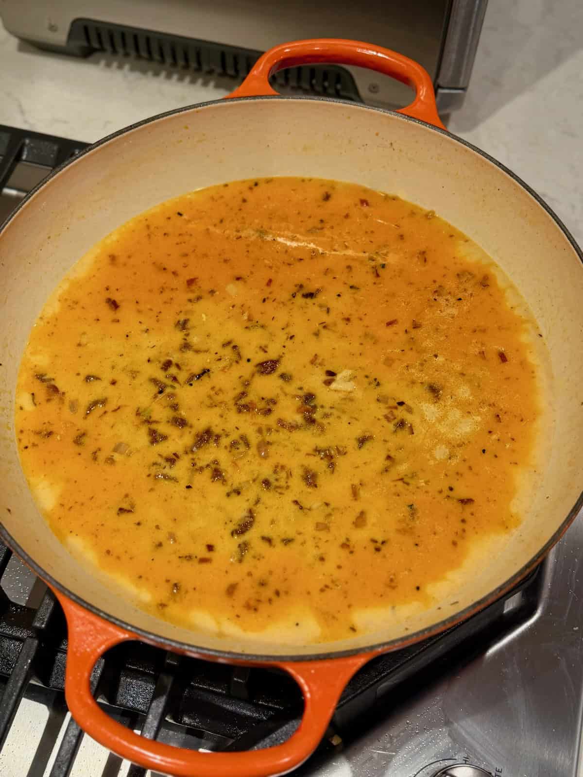 Broth and cream simmer with garlic, shallots, and sun-dried tomatoes in a pan.
