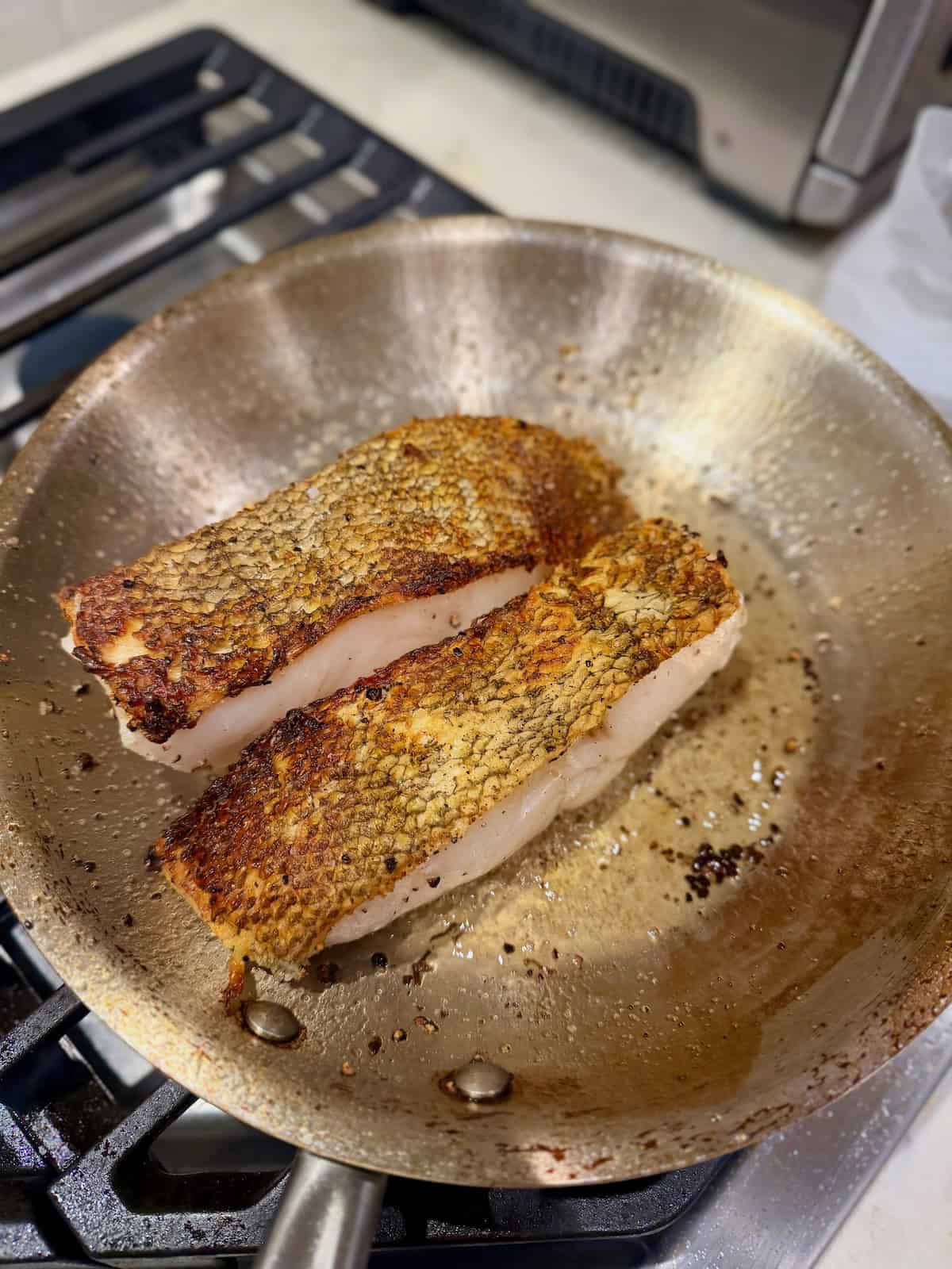 Two Chilean sea bass fillets cook with their golden, crispy skin facing up.