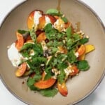 A tan bowl with sliced peacharines, cheese, fresh herbs, and pine nuts
