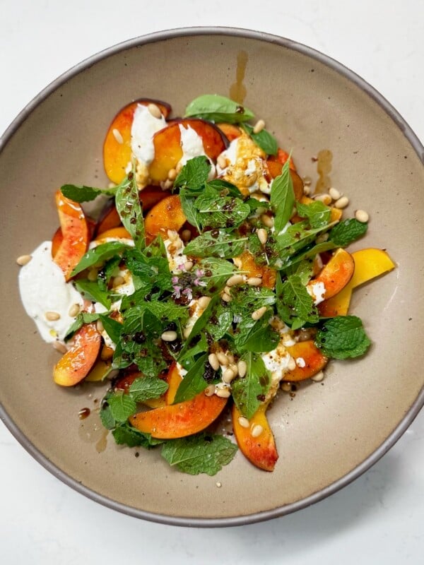 A tan bowl with sliced peacharines, cheese, fresh herbs, and pine nuts