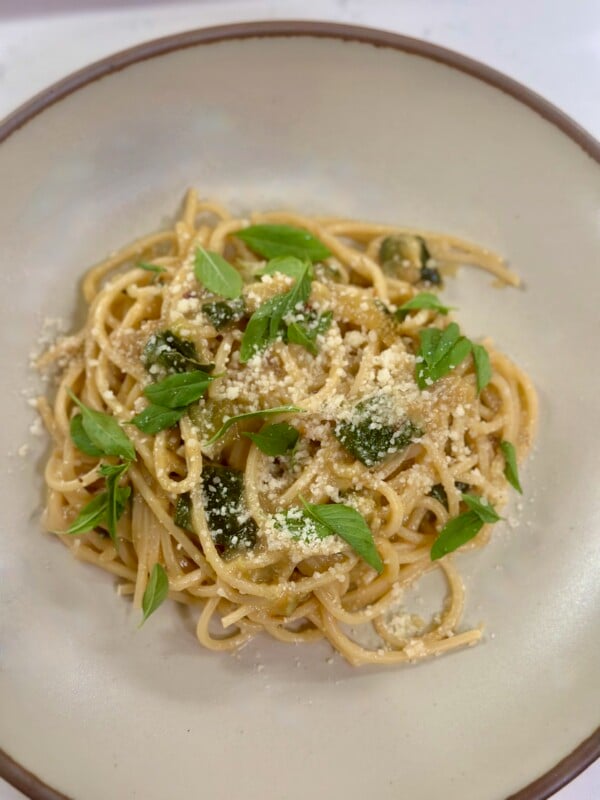 A white bowl with spaghetti tossed in a zucchini sauce topped with cheese and basil