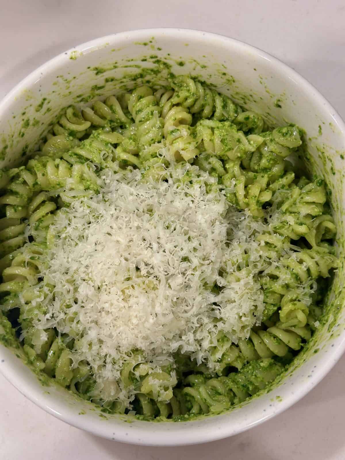 A white bowl with fusili pasta, green pesto, and grated parmesan cheese