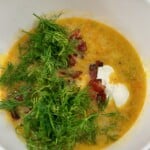 A white bowl with dill pickle soup, fresh dill, sour cream, and bacon