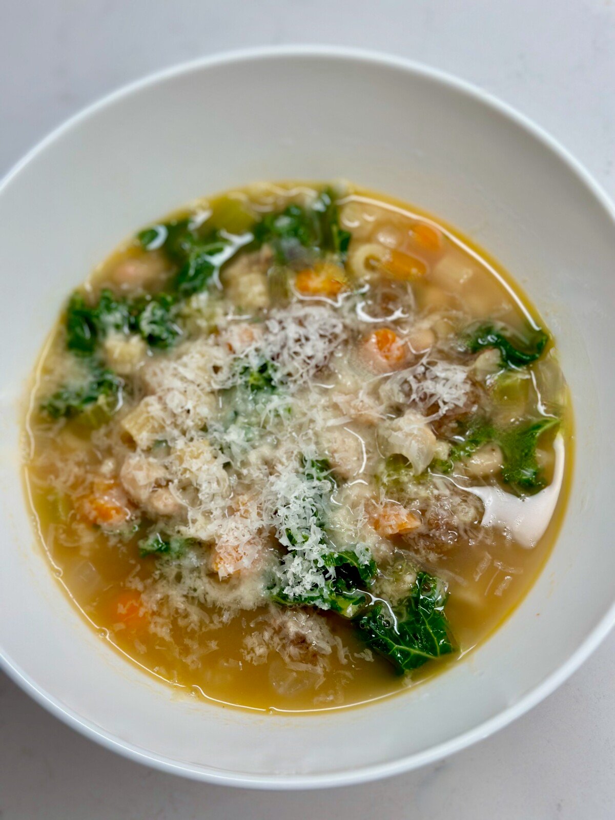 White bowl of soup with beans, pasta, kale, carrots, and parmesan cheese