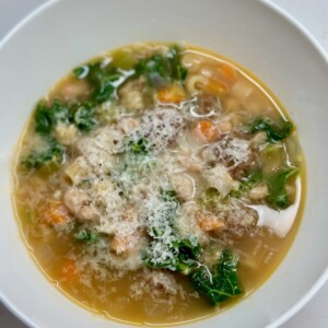 White bowl of soup with beans, pasta, kale, carrots, and parmesan cheese