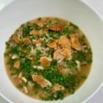 A white bowl with chicken soup, chives, crispy chicken skin, and herb sauce