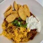 A white bowl with chili topped with sour cream, shredded cheddar cheese, sliced green onions, and fritos.