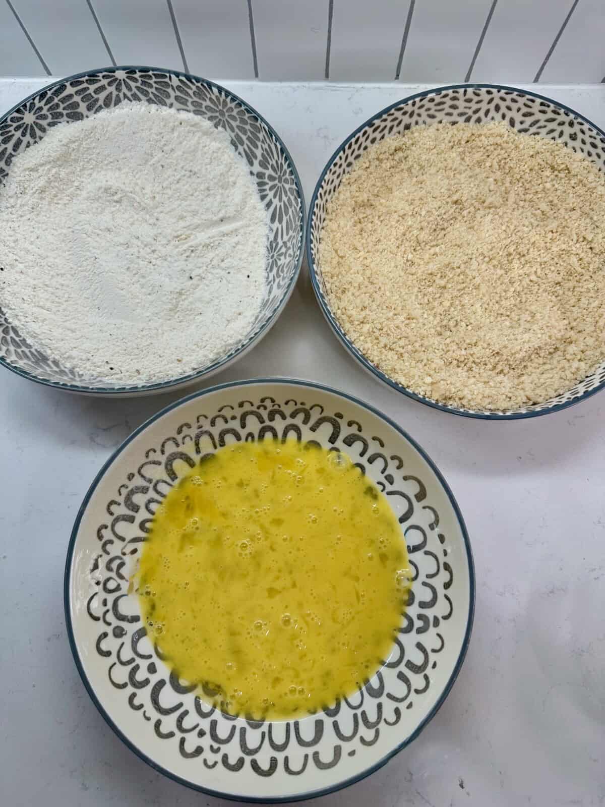 Three shallow bowls showing the breading station with flour, whisked eggs, and panko–Parmesan mixture.