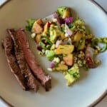 A serving plate with three strips of grilled skirt steak next to a scoop of peach, corn, and avocado salsa