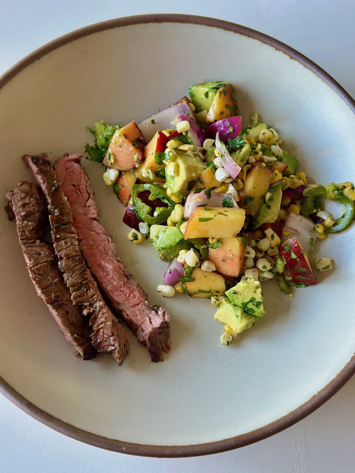 A serving plate with three strips of grilled skirt steak next to a scoop of peach, corn, and avocado salsa