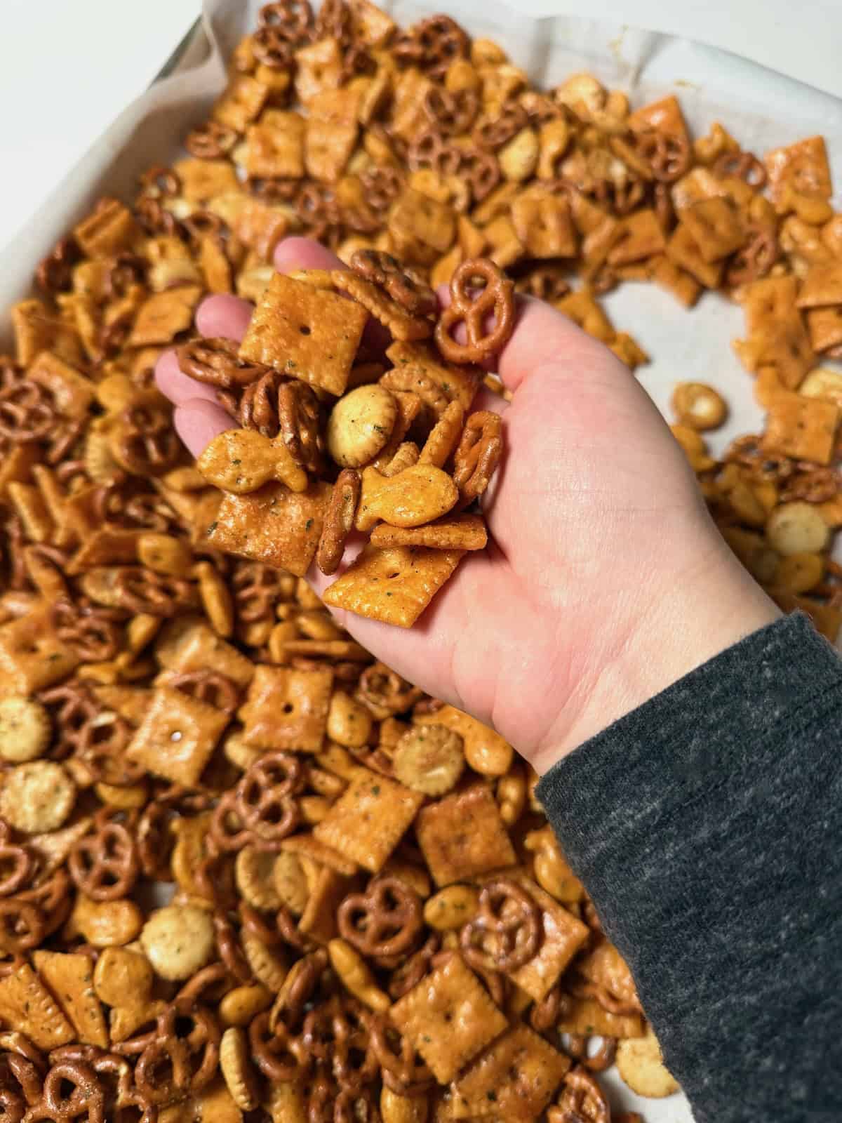 Hand holding baked Cheez-It ranch snack mix with pretzels, Goldfish, and oyster crackers.