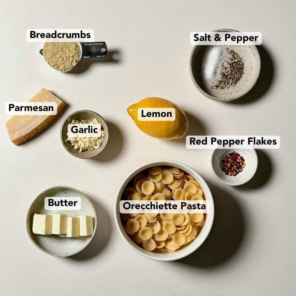 Ingredients for lemon garlic orecchiette including orecchiette pasta, lemon, garlic, butter, parmesan, and breadcrumbs arranged on a white surface.