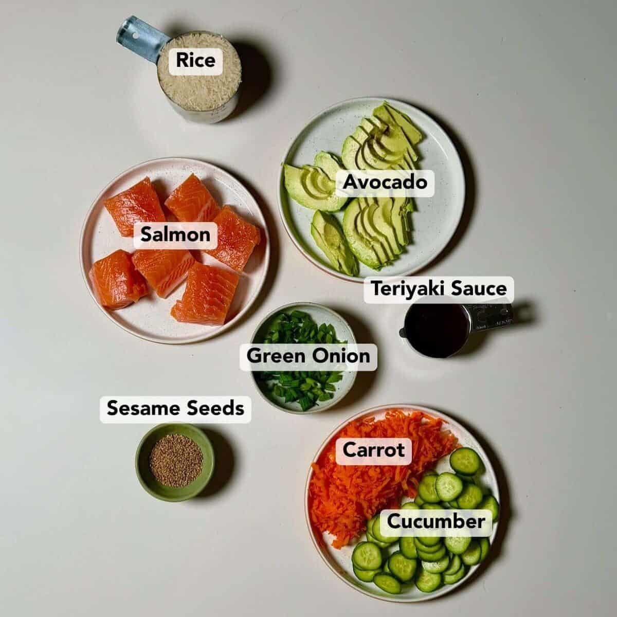 A variety of ingredients for a teriyaki salmon bowl including salmon, avocado, cucumber, rice, and sauce arranged on a white surface.