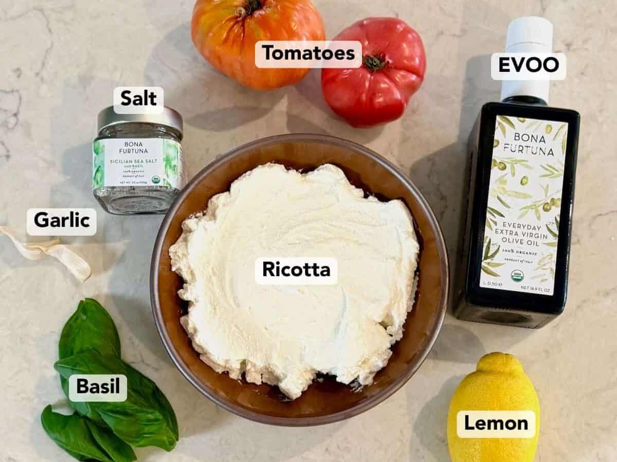 Ingredients for whipped ricotta bruschetta dip including ricotta, tomatoes, basil, lemon, garlic, olive oil, and salt.