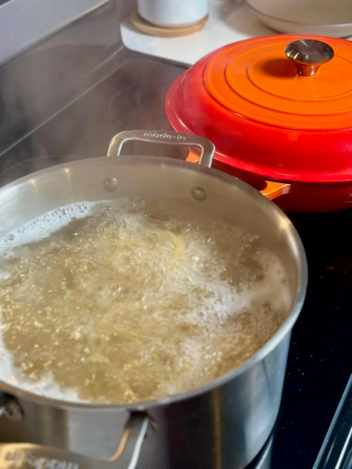 A large pot with pasta cooking in boiling water with an orange braiser next to it.
