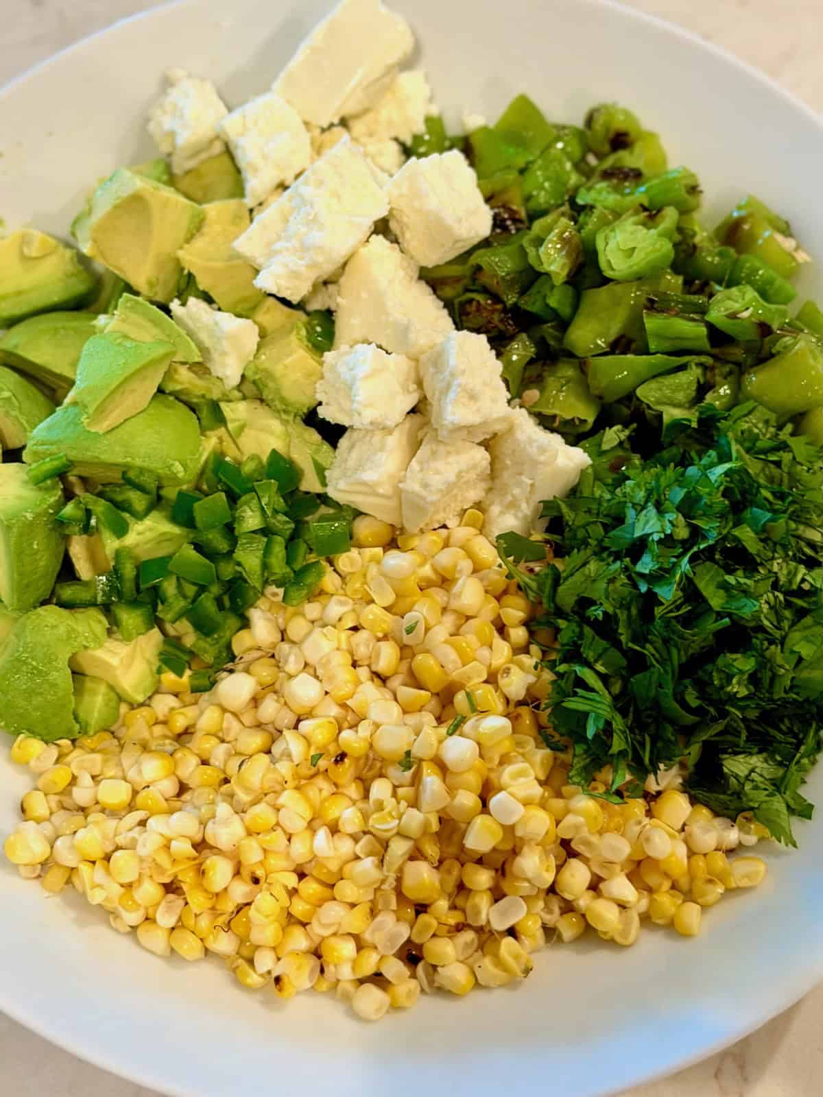 Chopped corn, avocado, cilantro, queso fresco, and shishito peppers in a bowl before mixing.