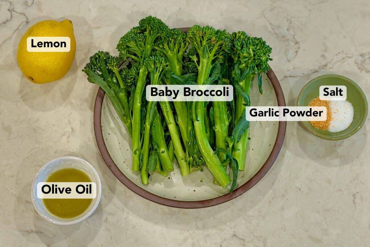 A plate of baby broccoli sits beside olive oil, lemon, salt, and pepper.