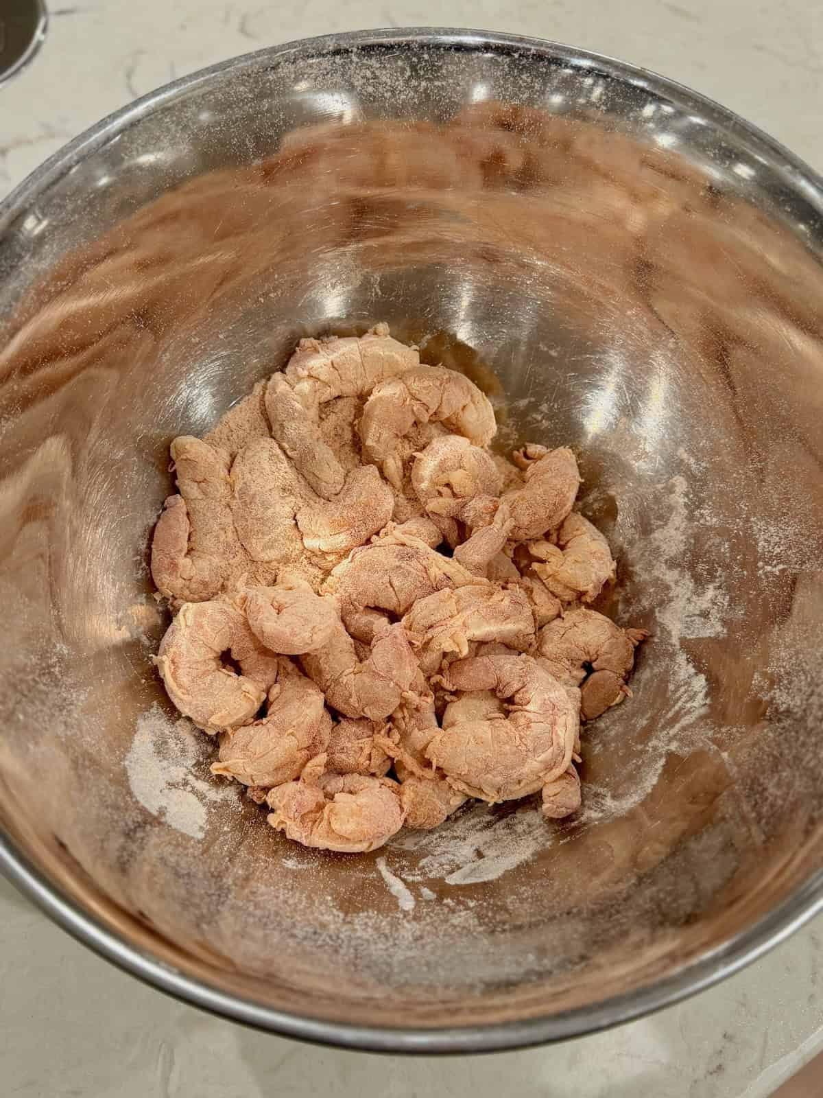 Raw shrimp are coated with flour and paprika in a large mixing bowl.