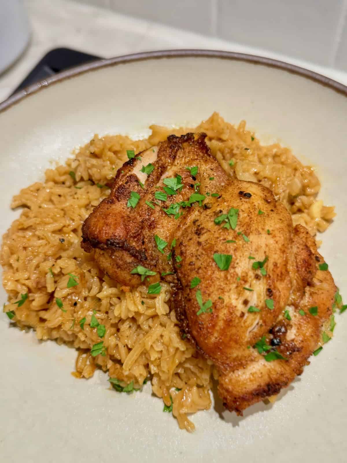 Golden brown chicken thigh rests on seasoned rice with chopped parsley.