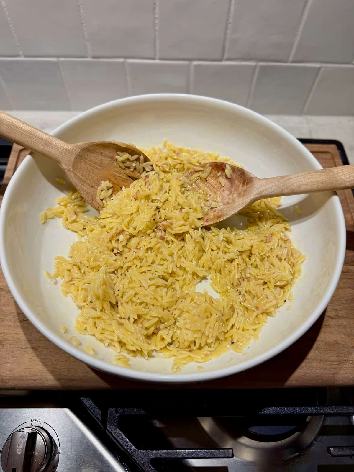 Cooked orzo sits in a large white mixing bowl with wooden serving spoons as the salad begins to be assembled.