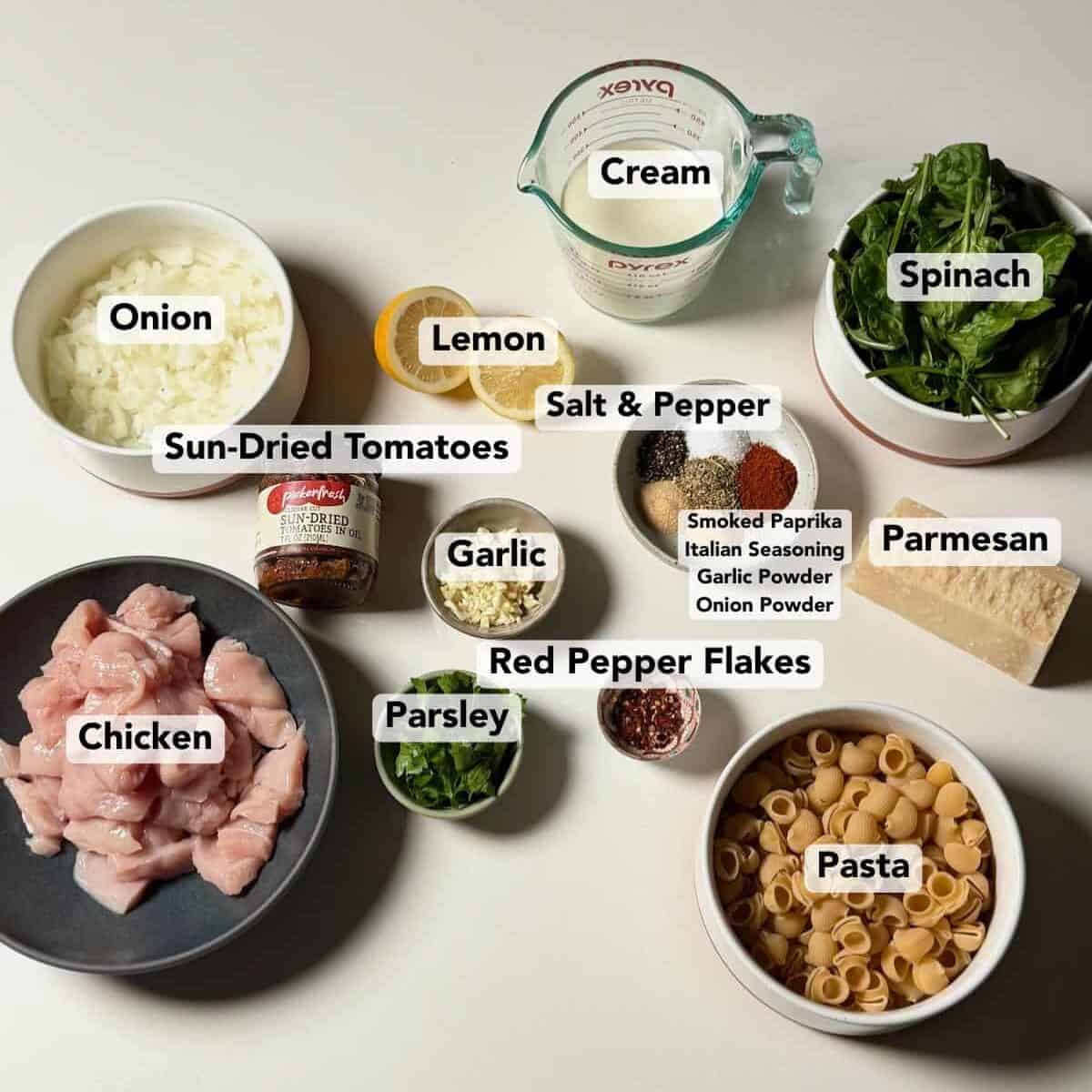 Ingredients for creamy sun-dried tomato chicken pasta arranged on a countertop, including chicken, pasta, sun-dried tomatoes, cream, spinach, and seasonings.