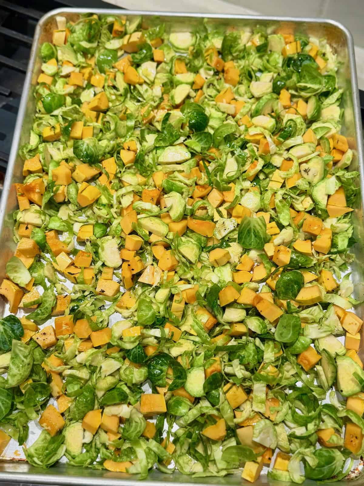 Diced delicata squash and shaved Brussels sprouts are spread across a baking sheet with olive oil and seasonings, ready to roast.