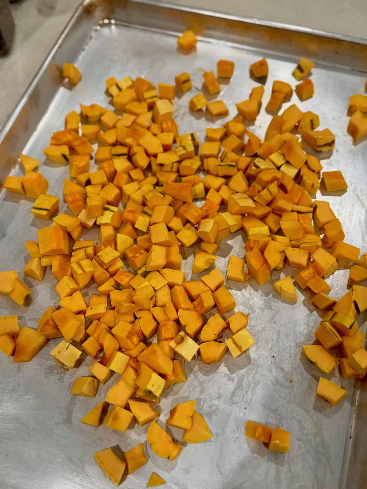 Small cubes of yellow and orange delicata squash with their skins attached are spread across a metal baking sheet.