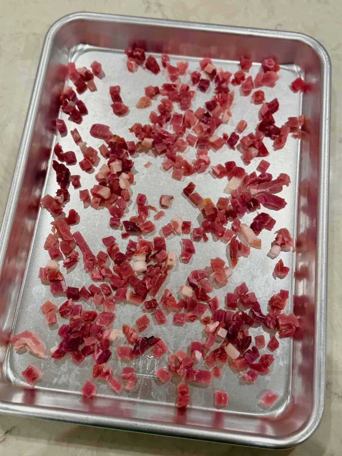 Small pieces of raw diced pancetta are spread across a baking sheet before cooking.