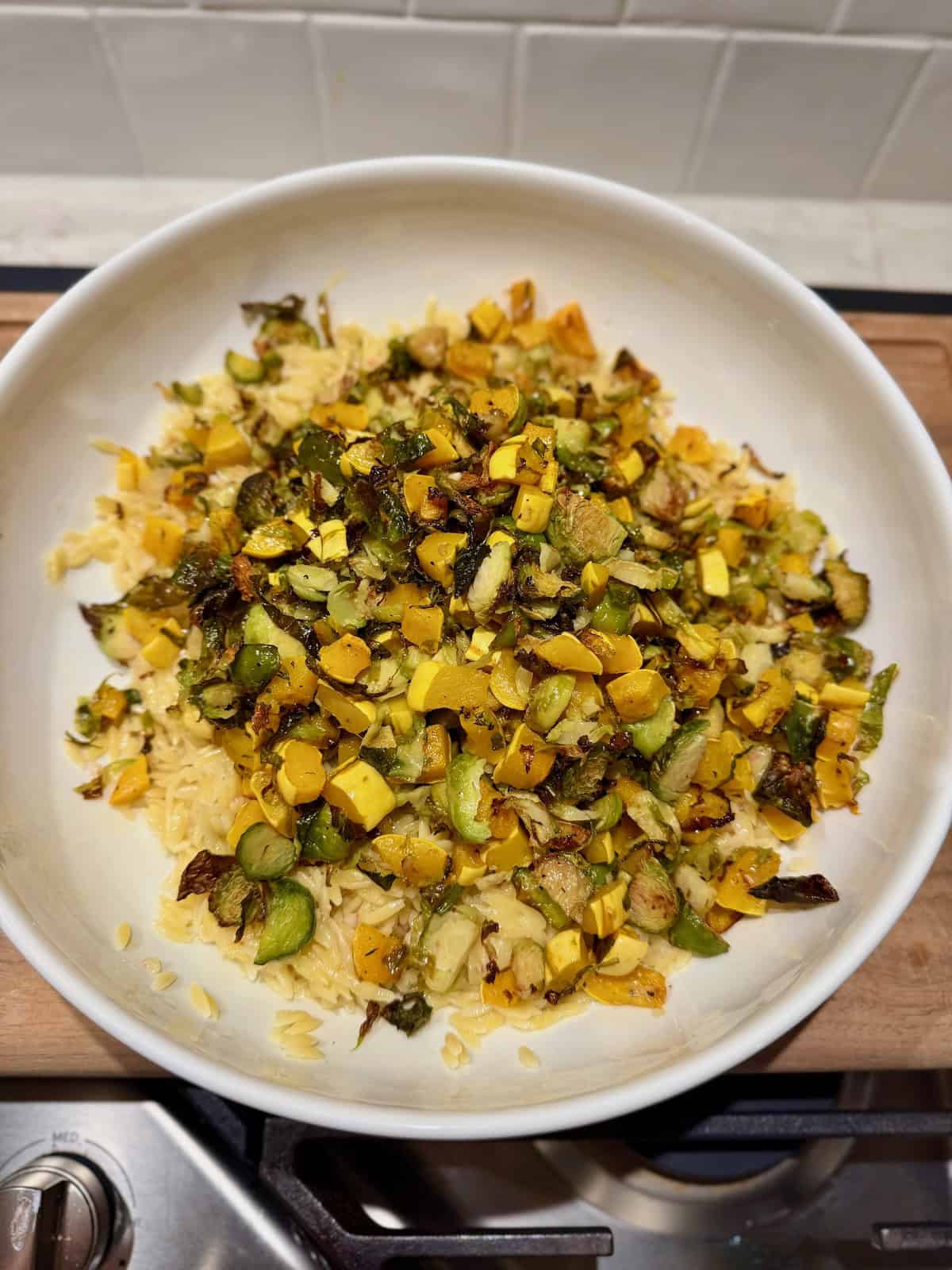 A large white bowl contains cooked orzo mixed with roasted delicata squash and Brussels sprouts before the remaining salad ingredients are added.