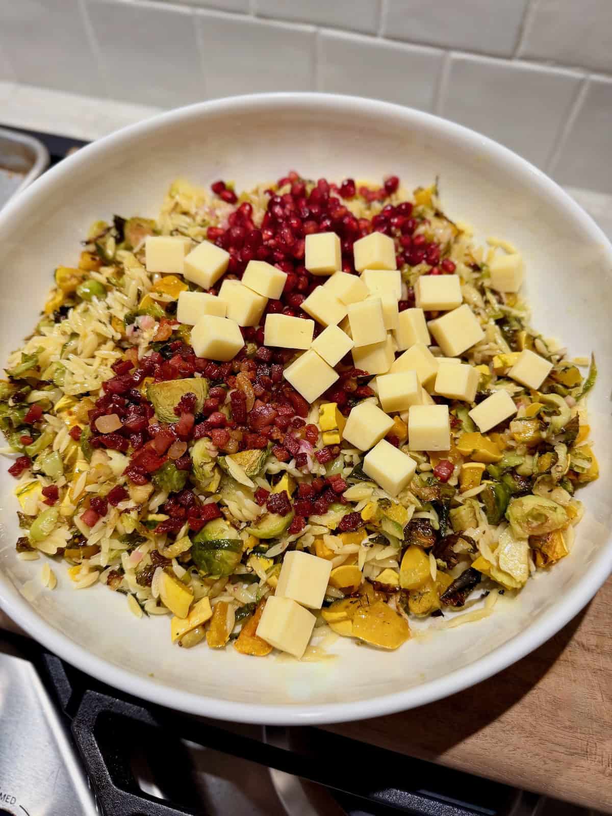 A white serving bowl holds orzo, roasted vegetables, cubed white cheddar, crispy pancetta, and pomegranate seeds before the salad is fully tossed together.