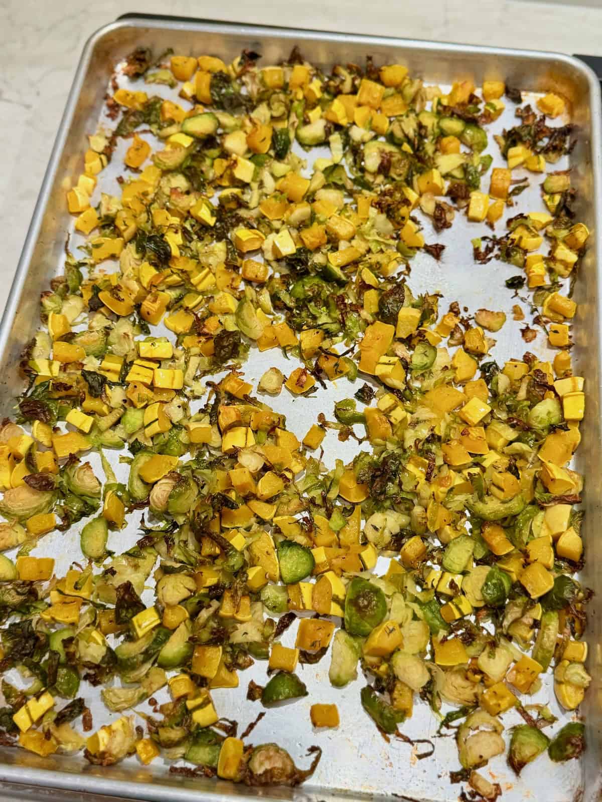 Roasted delicata squash and Brussels sprouts are spread across a metal baking sheet, with golden squash pieces and browned, crispy edges on the vegetables.
