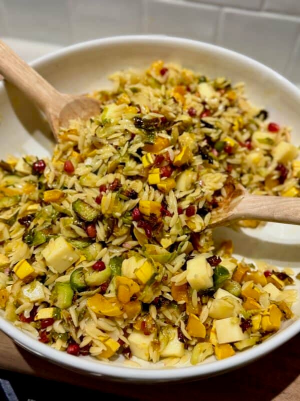 A white serving bowl filled with warm orzo salad, roasted delicata squash, Brussels sprouts, white cheddar, pancetta, and pomegranate seeds, with wooden spoons lifting the salad.