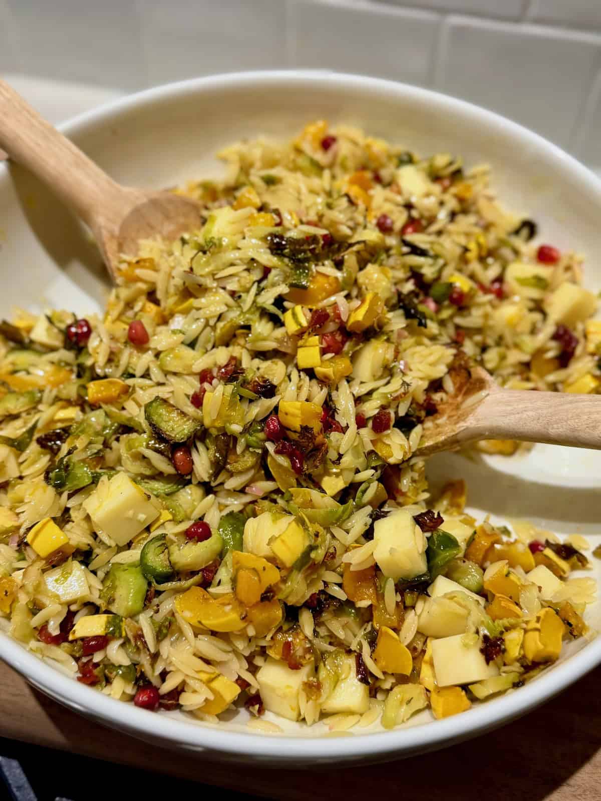 A white serving bowl filled with warm orzo salad, roasted delicata squash, Brussels sprouts, white cheddar, pancetta, and pomegranate seeds, with wooden spoons lifting the salad.