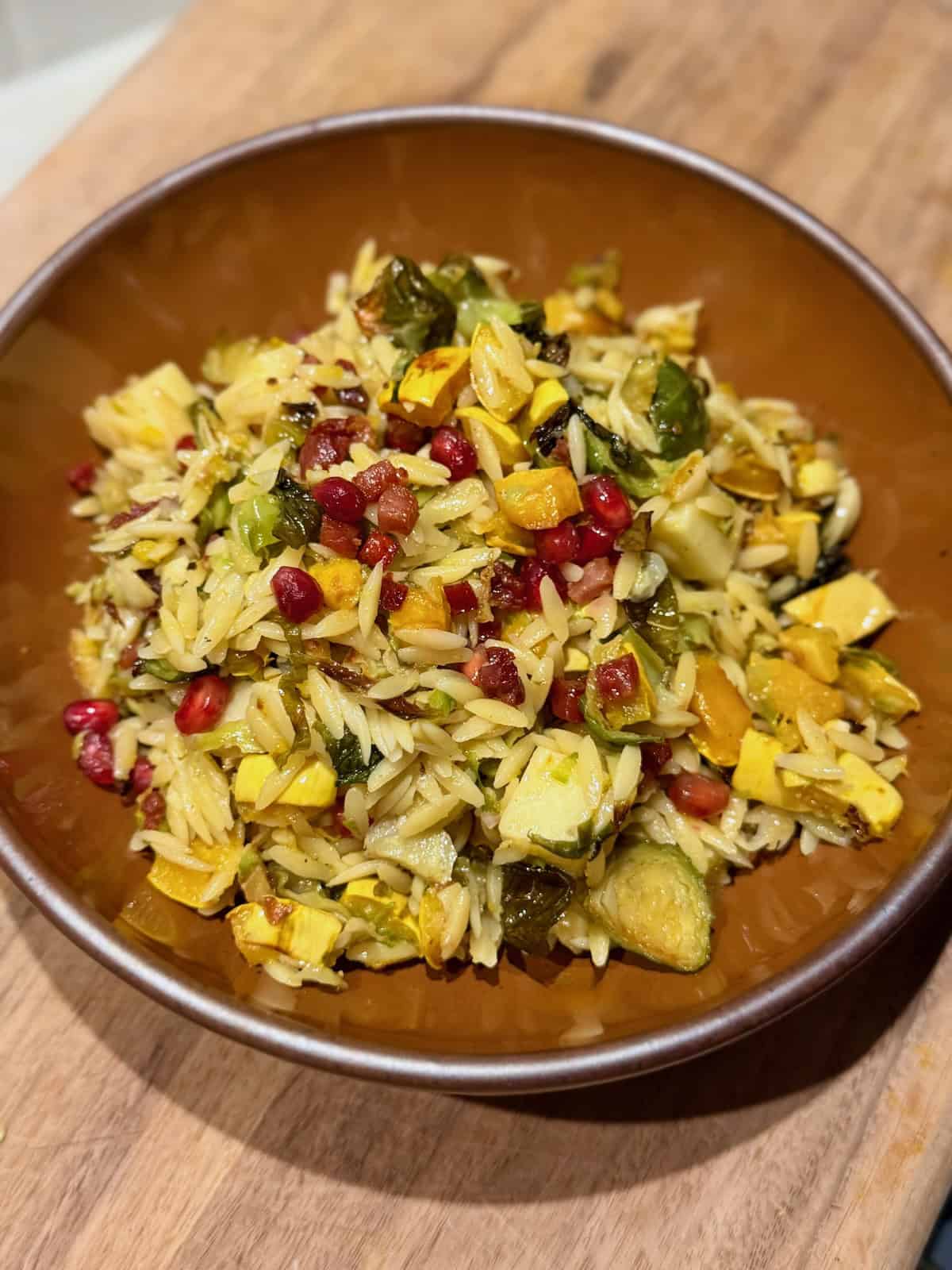 A brown ceramic bowl filled with warm orzo salad, roasted delicata squash, Brussels sprouts, white cheddar cubes, crispy pancetta, and pomegranate seeds.